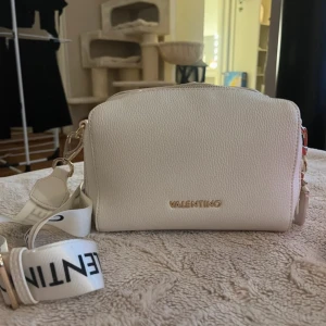 Vit axelväska från Mario Valentino - Snygg vit axelväska från Mario Valentino i strukturerat skinnimitation. Väskan har bred axelrem med logga, guldfärgade detaljer och dragkedja upptill. Embossad logga på ena sidan och praktisk ficka på baksidan. Perfekt storlek för det viktigaste.