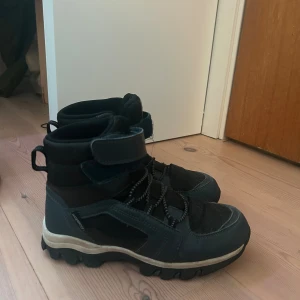 Svarta vinterkängor från Pax - Svarta kängor från Pax med grov sula, snörning och kardborreband upptill. Skorna har detaljer i syntet och textil, samt reflexdetaljer. Perfekta för kalla dagar med sin robusta design och höga skaft.