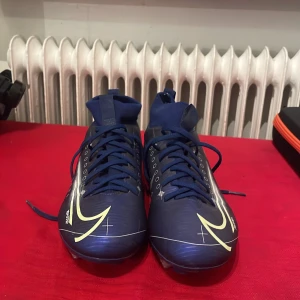 Blå Nike fotbollsskor med snörning - Säljer ett par blå Nike fotbollsskor med högt skaft och snörning. Skorna har en stor gul Nike-logga på sidan och metall-dobbar för bästa grepp på planen. Tillverkade i syntetmaterial och designade för att ge bra stöd och känsla vid spel.