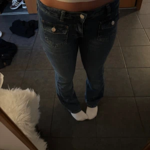 Blå jeans med fickdetaljer - Säljer ett par blå jeans med snygga fickor framtill med lock och silverknappar. Jeansen har en klassisk rak passform och normal midja. Perfekta för dig som gillar en enkel men ändå cool stil.