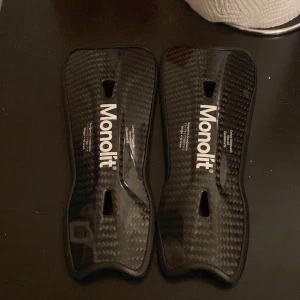 Svarta benskydd från Monolit - Säljer ett par svarta benskydd från Monolit i kolfiber-look. De är lätta, smidiga och har en sportig design med vit logga. Perfekta för dig som vill ha extra skydd på planen utan att kompromissa med stilen.