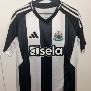 Newcastle United adidas matchtröja M - Snygg Newcastle United fotbollströja från adidas i slim fit. Klassisk svartvit randig design med klubbmärke, sponsortryck och gula detaljer på ärmen. Tillverkad i lätt och ventilerande AEROREADY-material, perfekt för match eller träning.