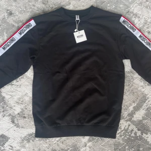 Svart dress från Moschino XS - Svarta mjukisbyxor från Moschino med vita och röda sidoremsor där Moschino-loggan repeteras längs benen. Byxorna har snörning i midjan och är tillverkade i mjukt bomullsmaterial. Perfekta för en chill och sportig look. Aldrig änvända prislappen är kvar på ny pris 3700 