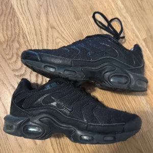 Svarta Nike sneakers med mesh och gummisula - Säljer ett par helsvarta Nike sneakers med mesh- och syntetöverdel, tydliga vågformade detaljer och robust gummisula med synliga luftbubblor. Klassisk Nike Swoosh på sidan och snörning framtill. Perfekta för dig som gillar sportig och urban stil.