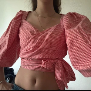 Rutig omlottopp med puffärm - Superfin röd och vit rutig omlottopp med stora puffärmar och knyt i midjan. Toppen har v-ringning och croppad passform, perfekt för en trendig look. 