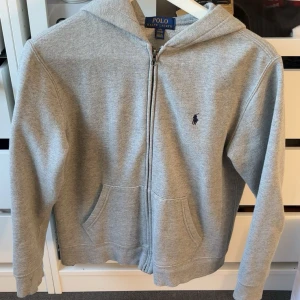 Grå zip hoodie från Polo Ralph Lauren - Klassisk grå hoodie från Polo Ralph Lauren med dragkedja framtill, huva och två fickor. Broderad logga på bröstet och mjukt material i bomullsmix. Perfekt för chill dagar och enkel att matcha med jeans eller joggers.