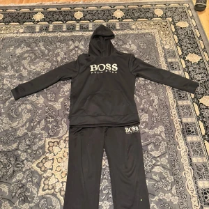 Svart hoodie från Hugo Boss - Svart hoodie från Hugo Boss med stor vit logga på bröstet. Tröjan har huva med snörning och en stor magficka framtill. Långärmad och i mjukt material, perfekt för chill dagar. Klassisk och stilren design som funkar året runt.