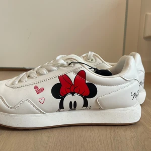 Vita sneakers Minnie Mouse Lefties - Snygga vita sneakers med Minnie Mouse-tryck på sidan och röd rosett. Skorna har snörning, platt sula och detaljer med hjärtan. Mjuk insida med röd innersula och text. Perfekta för dig som gillar Disney och vill sticka ut med en lekfull stil.
