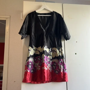 Blommig omlottklänning från Vero Moda - Kort omlottklänning från Vero Moda i svart, rött och vitt med blommigt mönster och v-ringning. Klänningen har korta ärmar och är tillverkad i ett glansigt, lätt material som ger en snygg siluett. Perfekt för dig som gillar färg och mönster!