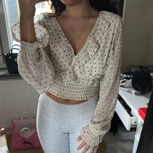 Prickig omlottblus med volangdetaljer - Supersöt beige omlottblus med svarta prickar, v-ringning och volangdetaljer längs halsringningen. Blusen är croppad med smockad midja och har långa, luftiga ärmar med resår vid handlederna. Perfekt för dig som gillar romantisk och trendig stil.