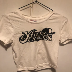 Vit croppad t-shirt Xtreme Clothing - Säljer en vit croppad t-shirt från Xtreme Clothing med svart tryck framtill. Trycket har cool text och en döskalle med keps. T-shirten har korta ärmar, rund halsringning och är i mjuk bomull. Perfekt för dig som gillar streetstyle och vill sticka ut. Storleken e x-small