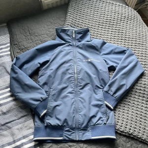 Blå vindjacka från Sail Racing - Blå vindjacka från Sail Racing med vit dragkedja och logga på bröstet samt nacken. Jackan har ribbade muddar med vit rand, ståkrage och två sidofickor. Materialet är lätt och syntetiskt, perfekt för blåsiga dagar. Snygg och sportig design med marinblå ton.