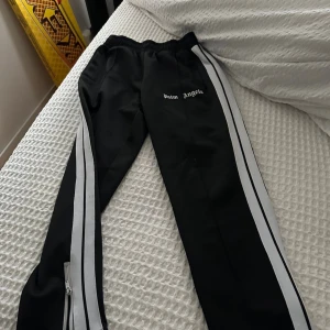 Palm Angels svarta trackpants med ränder - Svarta trackpants från Palm Angels med vita dubbla ränder längs sidorna och broderad logga på låret. Byxorna har elastisk midja och dragkedjor vid bensluten för extra stilpoäng. Perfekta för en sportig och trendig look.
