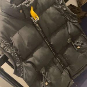 Svart dunväst från Parajumpers - Svart dunväst från Parajumpers med klassisk gul detalj vid kragen och broderad logga på ryggen. Västen har dragkedja framtill, två fickor med dragkedja och coola metalldetaljer. Perfekt för lager-på-lager och streetstyle.