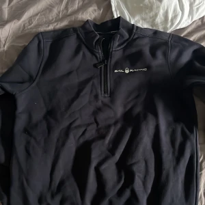 Svart Sail Racing half zip tröja - Svart långärmad tröja från Sail Racing med half zip och ståkrage. Tröjan har en stilren logga på bröstet och en mindre logga i nacken. Materialet känns mjukt och sportigt, perfekt för dig som gillar en clean look.