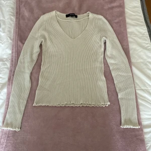 Ribbad vit långärmad tröja från Vero Moda - Supersnygg ribbad vit långärmad tröja från Vero Moda med v-ringning och vågiga kanter vid ärmslut och nederkant. Perfekt att styla med jeans eller kjol för en clean look. Mjuk och stretchig, sitter tajt men bekvämt.