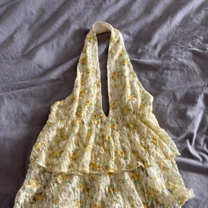 Blommig spetsig halterneck topp Gina Tricot - Supersöt blommig halterneck topp från Gina Tricot i ljusgul spets med volanger. Djup v-ringning framtill och öppen rygg. Toppens blommönster går i gult och grönt på en vit bas. Perfekt för sommarens alla tillfällen! Storlek S