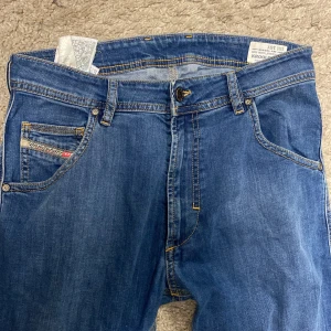 Blå Diesel Krooley JP jeans - Snygga blå jeans från Diesel, modell Krooley JP. De har klassisk femficksdesign, regular slim carrot-passform och coola detaljer som Diesel-logga på myntfickan och läderpatch bak. Jeansen är tillverkade i stretchigt denim för extra komfort.