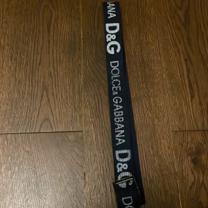 Svart Dolce & Gabbana bälte med logga - Snyggt svart bälte från Dolce & Gabbana med tydlig vit D&G-logga och text längs hela bandet. Bältet har en klassisk metallspänne och är gjort i ett slitstarkt syntetmaterial. Perfekt accessoar för att lyfta din outfit med en lyxig touch.