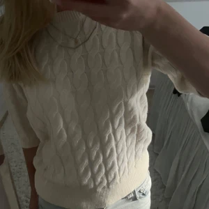 Krämvit stickad tröja från AWARE - Säljer en krämvit stickad tröja från AWARE i storlek XS. Tröjan har ett snyggt kabelstickat mönster, rund hals och korta ärmar. Perfekt att matcha med jeans eller kjol för en chill och stilren look.