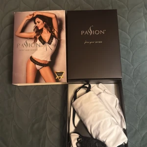 Vit och svart linnetopp från Passion - Säljer en snygg ny vit linnetopp från Passion med svarta spetsdetaljer och smala axelband. Toppen har en v-ringning med svart spets och en liten rosett framtill. Materialet är mjukt och glansigt, perfekt för en chic look.