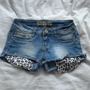 lågmidjade jeansshorts  - Snygga blå jeansshorts från Realia Luxury med eopardmönstrade fickor och guldiga paljettpatchar på bakfickorna. Shortsen har låg midja, klassiska fickor och en unik knapp framtill.  Storlek 26 ( xs) Midjemått: 35cm Grenlängd: 22cm