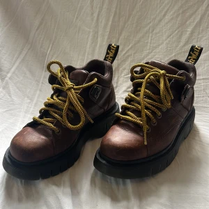 Brunt läder Dr Martens - Låga Dr Martens kängor i brunt läder med gula skosnören. Tjock sula med ”Soft Wair - comfort footbed”. Storlek 39. Gott skick, bara använda ett fåtal gånger.