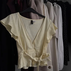 Ljusgul omlottopp från H&M - Supersöt ljusgul omlottopp från H&M med v-ringning, volangiga korta ärmar och knyt i midjan. Tunt, luftigt material som känns perfekt för varmare dagar. Enkel att styla med jeans eller kjol för en fräsch look. Storlek XXS. Finns en litet tråd men var där när jag köpte den
