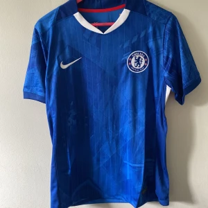Chelsea FC blå matchtröja Nike M - Chelsea FCs officiella matchtröja från Nike i blått med diskret mönster och klubbmärke på bröstet. Tröjan har vit detalj vid kragen och är tillverkad i lätt, ventilerande polyester. Perfekt för dig som vill visa ditt stöd för Chelsea på läktaren eller planen.