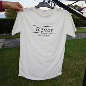 Vit t-shirt med tryck Rêver - Vit t-shirt med svart texttryck 'Rêver To dream Just be yourself' på bröstet. Klassisk rund hals och korta ärmar. T-shirten har en clean och enkel look, perfekt för dig som gillar stilrena plagg med budskap. Materialet känns mjukt och bekvämt mot huden.
