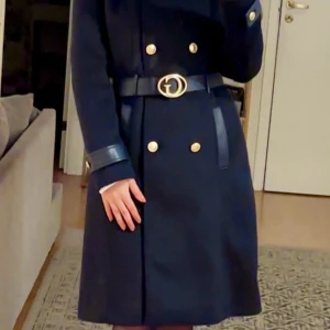 Mörkblå kappa från Guess med bälte - Snygg mörkblå kappa från Guess med guldiga knappar och ett stilrent bälte med metallspänne i midjan. Kappan har lång ärm och detaljer i skinnimitation vid ärmslut och bälte. Perfekt för dig som vill ha en klassisk och trendig look.