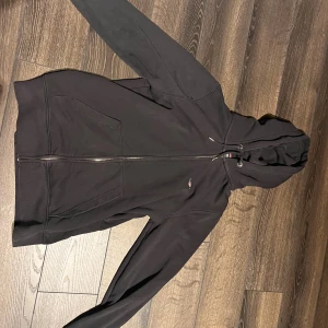 Svart hoodie från GANT med dragkedja - Svart hoodie från GANT i storlek S. Klassisk modell med huva och snörning, hel dragkedja framtill och två stora fickor. Tillverkad i mjuk bomullsmix, perfekt för chill dagar. Diskret GANT-logga på bröstet och snygga detaljer vid huvan. Enda felet är att ena snöret e skadat 