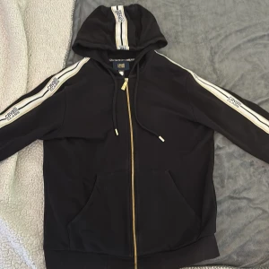 Svart hoodie Cavalli Class med guldzip - Svart hoodie från Cavalli Class med guldiga dragkedjor och vita/guldiga ränder med logga längs ärmar och huva. Klassisk känguruficka framtill, snörning i huvan och ribbade muddar. Snygg streetstil med exklusiv känsla.