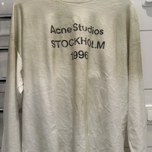 Vit långärmad tröja Acne Studios - Vit långärmad tröja från Acne Studios med svart tryck på bröstet där det står 'Acne Studios Stockholm 1996'. Tröjan har avslappnad passform och är gjord i mjuk bomull. Perfekt för en clean och stilren look.
