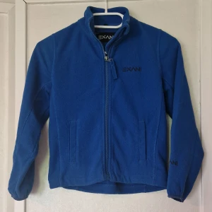 Blå fleecetröja från Exani - Blå fleecetröja från Exani med hel dragkedja framtill och två fickor. Jackan har hög krage, broderad logga på bröstet och ärmarna samt elastiska muddar vid ärmslut. Perfekt för kyliga dagar när du vill vara både varm och bekväm. Enda skavanken jag har hittat är slitage vid öppningen för händerna (se bild).