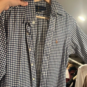 Rutig blåvit skjorta från Ralph Lauren - Säljer en slim fit stretch oxford-skjorta från Ralph Lauren i blåvitt rutigt mönster. Skjortan har klassisk krage, knappar framtill och den ikoniska broderade loggan på bröstet. Perfekt för dig som gillar stilrena och tidlösa plagg.
