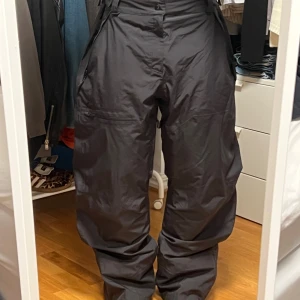 Oakley Oversize Skidbyxor i Gore-Tex - Svarta skidbyxor i Gore-Tex med bred passform och flera praktiska fickor, perfekta för parkråttan. Byxorna har hög midja, dragkedja och knapp framtill samt förstärkta partier vid bensluten. Använda under en skidvecka med lätt slitage från pjäxorna. Syns endast vid noggrann inspektion.