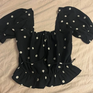 Svart prickig blus från H&M - Supersöt svart blus från H&M i storlek XS med vita prickar. Modellen har puffiga korta ärmar, smockad i midjan! ❤️!!!!välj postnord när du beställer inte instabox!!