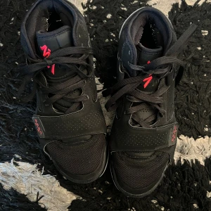 Nike Air Jordan Zion 2 black - Svarta Nike Zoom Air sneakers med högt skaft och grå detaljer på hälen. Skorna har snörning, kardborreband och röd Nike-logga på plösen. Ovandelen är i en mix av mesh och syntetmaterial med perforerade partier för extra stil.