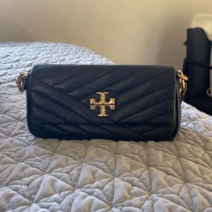 Svart quiltad axelväska Tory Burch - Snygg svart quiltad axelväska från Tory Burch med guldiga metalldetaljer och ikonisk logga framtill. Väskan är tillverkad i skinn och har ett stilrent, avlångt format med justerbar axelrem. Perfekt för dig som vill ha en trendig och klassisk accessoar.