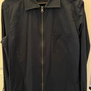Mörkblå overshirt med dragkedja - Säljer en mörkblå overshirt med klassisk krage, bröstficka och hel dragkedja framtill. Jackan har lång ärm och enkel, stilren design som passar till många outfits. Perfekt för lager-på-lager och snygg till både jeans och chinos.