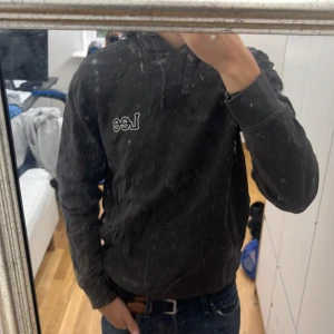 Svart hoodie från Lee med tryck - Svart hoodie från Lee med stort vitt logotryck på ryggen och mindre logga på bröstet. Klassisk huva med snörning och känguruficka framtill. Regular fit och mjukt material som känns skönt mot huden. Perfekt för chill dagar eller när du vill ha en enkel streetstyle.