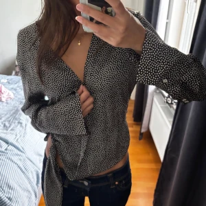 Prickig omlottblus från Zara XS - Svart omlottblus från Zara i storlek XS med vita prickar. Blusen har långa ärmar, knytband i midjan och v-ringad hals. Materialet är glansigt och lätt, vilket ger en snygg och flowy känsla. Perfekt för dig som gillar trendiga och feminina detaljer.