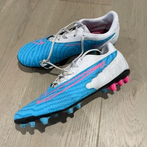 Nike blå fotbollsskor med rosa detaljer - Säljer ett par Nike fotbollsskor i blått med vita och rosa detaljer. Skorna har snörning, platt sula med dobbar i blått och rosa, samt ett sportigt mönster på ovandelen. Perfekta för dig som vill sticka ut på planen.