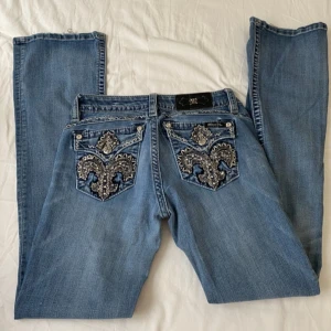 Blå Miss Me jeans med broderade fickor - Snygga blå jeans från Miss Me med bootcut-modell och detaljerade bakfickor med broderier och nitar. Jeansen har låg midja och klassisk femficksdesign. Perfekta för dig som gillar statement-jeans med coola detaljer.