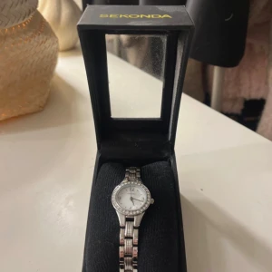 Sekonda silverfärgad armbandsklocka - Stilren armbandsklocka från Sekonda i silverfärg med klassisk rund urtavla och länkarmband. Urtavlan är vit med tydliga siffror och ramen är dekorerad med små stenar för extra bling. Klockan levereras i original ask.