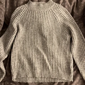 Grå stickad tröja från 157 - Säljer en chunky grå stickad tröja från 157 med grov struktur och ribbade muddar. Tröjan har högre hals och långa ärmar, perfekt för lager på lager under kyliga dagar. Materialmix av akryl, polyester och nylon för en mjuk och skön känsla. Färgen är mer rättvis på andra bilden