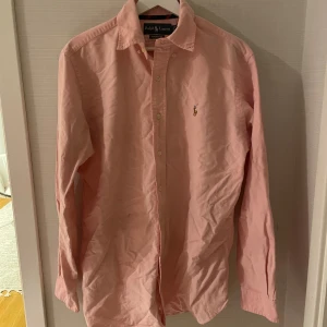 Rosa skjorta från Ralph Lauren M - Snygg klassisk rosa skjorta från Ralph Lauren i bomull med broderad logga på bröstet. Skjortan har lång ärm, knappar framtill och klassisk krage. Passformen är normal och materialet känns mjukt och skönt mot huden. Perfekt för dig som gillar stilrena plagg med ikonisk design.