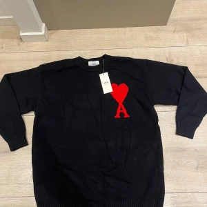 Svart stickad tröja från Ami - Svart stickad tröja från Ami Alexandre Mattiussi med rund hals och ribbade muddar. På bröstet finns ett stort rött hjärta och bokstaven A, vilket ger en unik och trendig look. Perfekt för dig som vill sticka ut med en snygg detalj.