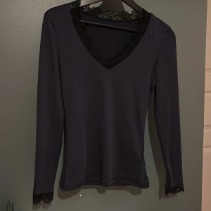 Mörkblå långärmad topp med spets - Snygg mörkblå långärmad topp med v-ringning och dekorativ svart spets runt halsringningen och ärmsluten. Toppens material känns mjukt och har en lätt stretch. 